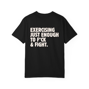 May include: Black t-shirt with the text "EXERCISING JUST ENOUGH TO F*CK & FIGHT" in a bold, white font. The shirt is a classic crew neck style, perfect for casual wear. The text is centered on the back of the shirt.