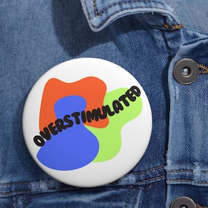 May include: A white button pin with the word "OVERSTIMULATED" in black, angled text. The pin features abstract shapes in orange, blue, and green. The pin is resting on a denim jacket.