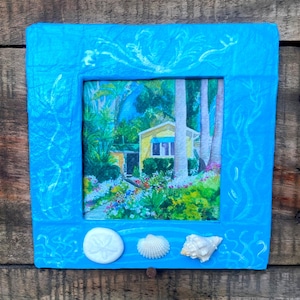 May include: A framed painting of a yellow house surrounded by lush greenery and colorful flowers. The painting is set within a bright blue frame adorned with white decorative accents and seashells.