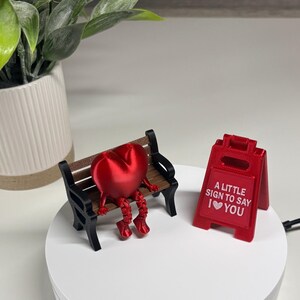 May include: A red heart-shaped figure with arms and legs sits on a miniature park bench. A red sign next to the bench reads "A LITTLE SIGN TO SAY I ❤️ YOU". The bench has black supports and brown slats.