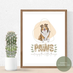 Op de afbeelding: Een ingelijste print met een hondenillustratie in bruin, wit en beige. Het kunstwerk bevat de tekst "PAWS" met pootafdrukken en de zin "and enjoy the little things". Een digitale downloadgrafiek staat in de rechter benedenhoek.