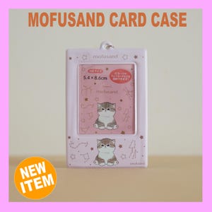以下が含まれることがあります： 上部に「MOFUSAND CARD CASE」の文字が入った薄いピンク色のカードケース。ケースには、ピンク色の背景、猫のイラスト、そして「5.4 x 8.6cm」の文字が入った長方形の窓があります。「NEW ITEM」ステッカーが左下にあります。