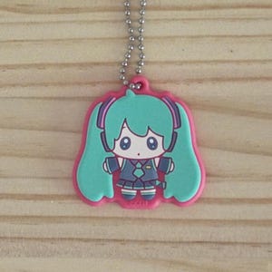 May include: A pink and teal cartoon character keychain with a silver ball chain. The character has teal hair, a blue and black outfit, and large eyes. The keychain is on a wooden surface.