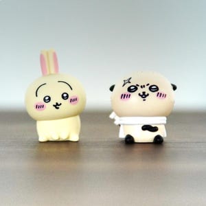 Könnte beinhalten: Zwei kleine Figuren im Cartoon-Stil. Eine ist ein hellgelbes Kaninchen mit rosa Ohren und rosigen Wangen. Die andere ist eine beigefarbene Dogge mit einem weißen Schal und schwarzen Akzenten. Beide haben ausdrucksstarke Gesichter.