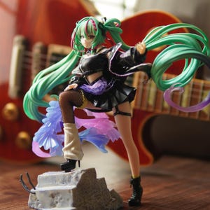 May include: A figurine of a character with long, flowing mint green hair, wearing a black cropped top, a black pleated skirt, fishnet stockings, and black boots. The character stands on a gray stone-like base with purple and blue fabric accents.