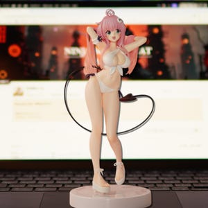 May include: A figurine of a female anime character in a white bikini, with pink hair and green eyes. The figure is standing on a white base, with a black loop behind it. The background is a computer screen.