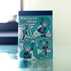 Puede incluir: Un bloc de notas turquesa y blanco con ilustraciones de un personaje de dibujos animados con largas coletas turquesas, que lleva un atuendo negro y auriculares. El bloc de notas tiene el texto "Hatsune Miku" y otro texto japonés.