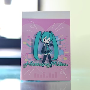 Puede incluir: Un bloc de notas rosa con una ilustración de dibujos animados de un personaje con largas coletas turquesas, que lleva un atuendo negro y auriculares. El bloc de notas tiene el texto "Hatsune Miku" en verde.