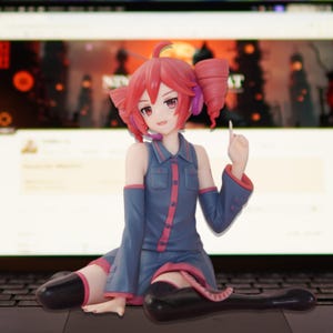 Pode incluir: Estatueta de uma personagem feminina de anime com cabelo ruivo e roupa azul. A figura está sentada no teclado de um laptop, com uma tela visível ao fundo. A personagem está usando fones de ouvido e tem um microfone.