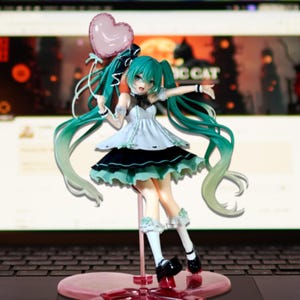 May include: A figurine of a female anime character with long, teal hair and a white and black dress. She holds a pink heart-shaped balloon. The figure is on a pink stand, with a laptop in the background.