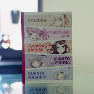 Könnte beinhalten: Ein farbenfrohes Notizbuch mit Anime-Illustrationen von weiblichen Charakteren. Das Notizbuch hat einen rosa Rücken und die Namen "DOLORIS", "MARUYAMA AYA", "TOYAMA KASUMI", "MINATO YUKINA" und "KURATA MASHIRO" sind auf dem Cover aufgedruckt.
