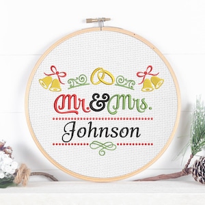 May include: A round, embroidered hoop art piece with the text "Mr. & Mrs. Johnson" in red, black, and green. The design includes wedding bells and rings. The hoop is made of wood and is displayed on a white surface.