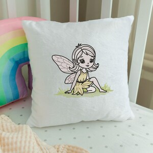 May include: A white, plush square pillow featuring an embroidered fairy design. The fairy has light skin, blonde hair, and wears a yellow dress. She is sitting on green grass with pink wings. A rainbow pillow is visible in the background.