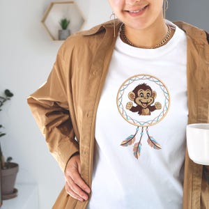 May include: White t-shirt featuring a cartoon monkey design within a dreamcatcher. The dreamcatcher has a gold border with blue and pink accents, and hanging feathers. The shirt is worn with a brown jacket.