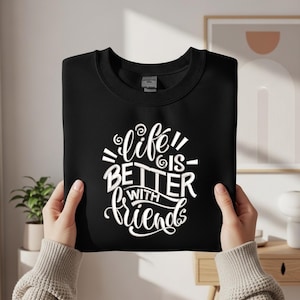 Könnte beinhalten: Schwarzes Sweatshirt mit dem weißen Text "Life is Better With Friends" in Schreibschrift. Das Sweatshirt wird von einer Person hochgehalten. Der Hintergrund umfasst eine Pflanze, einen Holztisch und ein gerahmtes Bild.