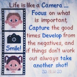 May include: Cross-stitch artwork with the text "Life is like a Camera." The design includes a film strip with cartoon faces, a camera, and the word "Smile!" The text continues with inspirational phrases in blue and red.