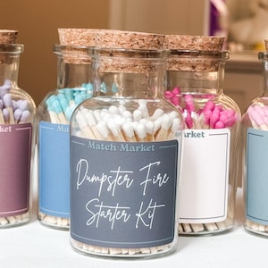 May include: Clear glass bottles filled with colored matches and cork stoppers. The front bottle has a label that reads "Match Market" and "Dumpster Fire Starter Kit" in white script. Other bottles contain blue, pink, and purple matches.