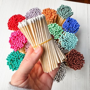 May include: A hand holds a bundle of wooden matches with white tips, secured by a yellow rubber band. Surrounding the bundle are several bouquets of matches with colorful heads in shades of red, pink, teal, green, orange, blue, purple, brown, and gray.