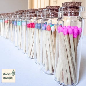 May include: A row of clear glass bottles with cork stoppers, each filled with wooden matches featuring vibrant, multi-colored heads in shades of pink, blue, orange, and white. The text Match Market is visible in the lower left corner of the image.