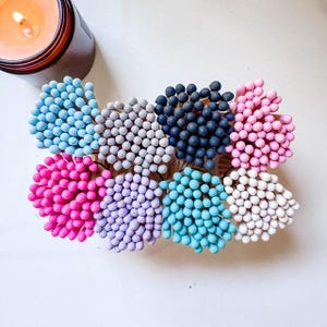 May include: A collection of colorful matches in various shades, including blue, pink, and gray, arranged in a circular pattern. A lit candle in a dark brown jar is in the upper left corner. The matches have a spherical head.