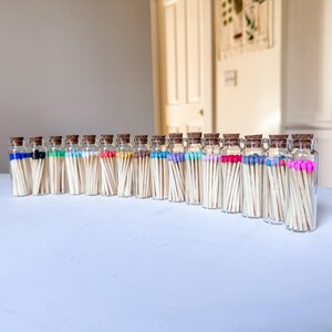 May include: A collection of clear glass vials arranged in a row, each containing wooden matches with colored tips. The vials are sealed with cork stoppers. The match tips display a variety of colors including blue, green, white, red, and pink.