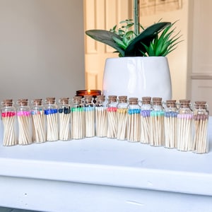 May include: A collection of seventeen small, clear glass bottles arranged in a row, each filled with wooden matches featuring different colored tips including pink, blue, black, green, white, yellow, purple, and brown, all sealed with cork stoppers.