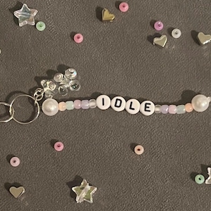 May include: A beaded keychain with the word "IDLE" spelled out in white letter beads. The keychain features a silver clasp, a cluster of clear beads, and a mix of pastel-colored beads. Scattered around are small star and heart-shaped embellishments.