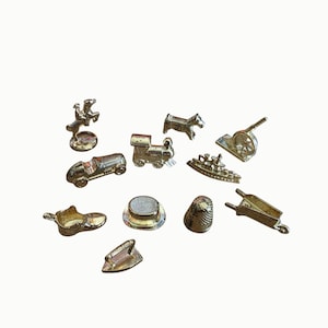 May include: A collection of ten small, metallic gold-toned game tokens featuring various shapes including a horse and rider, a dog, a cannon, a race car, a steam locomotive, a ship, a shoe, a top hat, a thimble, a wheelbarrow, and an iron.