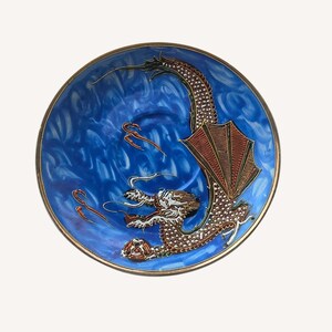 May include: A round decorative plate featuring a vibrant blue background with swirling patterns. A detailed dragon with textured scales, large wings, and a long tail is depicted in the center, holding a small circular object in its claw.