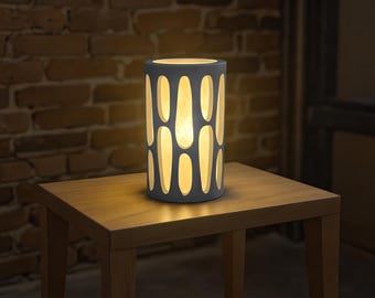 May include: A cylindrical gray lamp with a pattern of elongated cutouts, emitting a warm, soft light. The lamp sits on a wooden table, with a brick wall in the background. The lamp's design creates a decorative lighting effect.