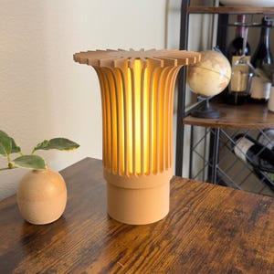 May include: A wooden table lamp with a unique design. The lamp has a cylindrical base and a top with a flower-like shape. The lamp emits a warm, inviting light. A small vase with a plant is on the left.