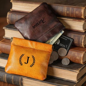 Puede incluir: Dos monederos de cuero, uno marrón y otro amarillo-anaranjado, descansan sobre una pila de libros antiguos. El monedero marrón está abierto, revelando efectivo y una tarjeta de crédito. El monedero amarillo-anaranjado tiene una "J" negra con una corona de laurel.