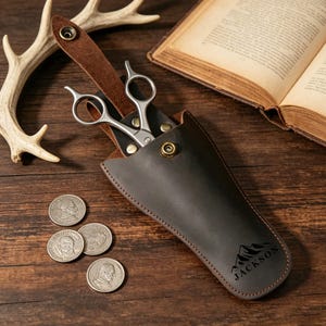 May include: A dark brown leather holster containing silver metal shears, secured with a brass snap button. The holster features a stamped mountain graphic and the text JACKSON. Three silver coins are positioned near the leather case.