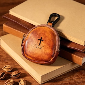 Pode incluir: Uma carteira de moedas de couro castanho com uma cruz preta e o nome "Eric" impresso na frente. A carteira tem fecho de correr e um mosquetão preto. A carteira está em cima de uma pilha de livros.