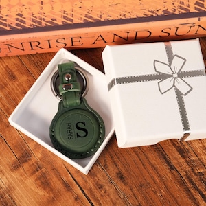 Puede incluir: Un llavero redondo de cuero verde oscuro con un anillo de metal y un cierre a presión. La superficie de cuero está grabada con el nombre SARAH y la letra S. El llavero se presenta dentro de una caja de regalo blanca con una cinta.