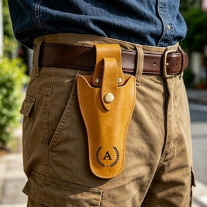Könnte beinhalten: Ein beigefarbenes Lederholster an einem braunen Gürtel mit einem vertikalen Riemen und einem metallischen Druckknopfverschluss. Das Holster zeigt ein schwarzes Emblem mit einem Lorbeerkranz, der den Großbuchstaben A in der Mitte umschließt.