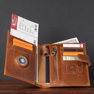 Puede incluir: Cartera de cuero marrón abierta con múltiples compartimentos para tarjetas, efectivo y documentos de viaje. Cuenta con un bolsillo con cremallera y cierre de botón. Incluye tarjetas de embarque y un AirTag de Apple. La marca "REIDD" es visible.