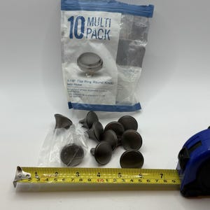 May include: A multi-pack of 10 satin nickel top ring round knobs. The knobs are dark gray and measure 1-1/8 inches in diameter. A measuring tape is next to the knobs.