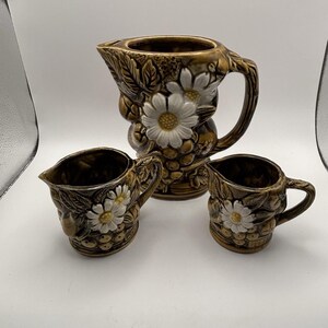 May include: A vintage ceramic pitcher and creamer set. The set features a brown glaze with embossed fruit, leaves, and white daisy accents. The pitcher has a large handle and spout, and the creamer has a smaller handle and spout.