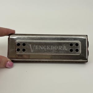 May include: A vintage harmonica with a silver metal casing. The word "Vencedora" is printed in the center. The harmonica has eight holes in two sets of four. The harmonica is held by a person's hand.