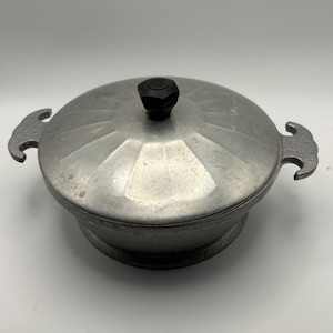 May include: A vintage aluminum casserole dish with a lid. The dish has a round shape with a textured surface and two handles on either side. The lid features a black knob and a geometric design. The dish is silver in color.