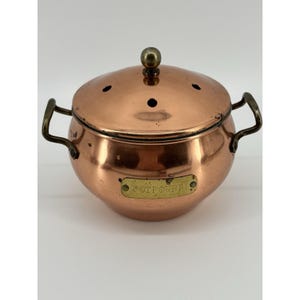May include: A copper potpourri pot with a lid and two handles. The pot has a shiny finish and a small brass label that reads "POTPOURRI". The lid has small holes for fragrance release and a brass knob.