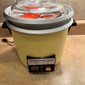 May include: A vintage yellow Hamilton Beach "The Pressureless Cooker" steamer and cooker. It has a clear glass lid, a white rim, and a black handle. The control panel has "Warm" and "Cook" settings.