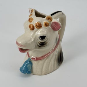May include: A ceramic cow creamer with a white base, brown spots, and a pink collar. The cow has a blue bell and a black and white handle. The creamer is designed to look like a cow's head.