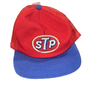 May include: A red and blue baseball cap with the STP logo. The cap features a red mesh back and a blue brim. The STP logo is white with a blue outline and is centered on the front of the cap.