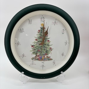 May include: A round Christmas-themed clock with a dark green frame and a white face. The clock face features a decorated Christmas tree with gifts and toys at the base. The clock hands are brown, and the numbers are marked around the edge.