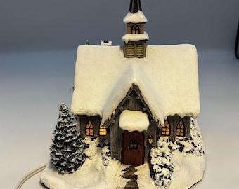 May include: A miniature illuminated church figurine featuring dark wood-textured walls, a brown arched door, and a steeple. The roof and base are covered in a thick layer of white snow, with a small evergreen tree and glowing yellow windows.
