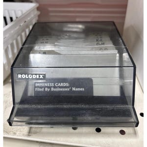 May include: A clear plastic Rolodex business card holder with a black base. The front of the holder has a label that reads "BUSINESS CARDS: Filed By Businesses' Names". The Rolodex logo is visible on the front.