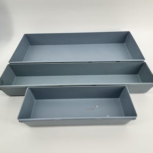 May include: Three gray rectangular plastic trays of varying sizes are stacked. The trays have a simple, minimalist design with straight sides and a flat bottom. The trays are empty and appear to be new.