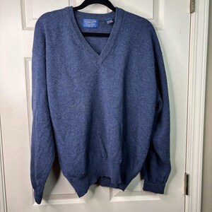 May include: A blue V-neck sweater with long sleeves. The sweater has a ribbed hem and cuffs. The brand name "Pendleton" is visible on a small tag near the neckline. The sweater is hanging on a black hanger against a white door.
